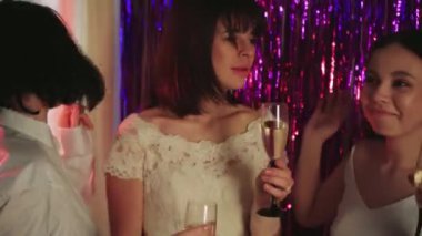 Klasik beyaz bir gelinlik giyen müstakbel gelin, bekarlığa veda partisini bir bardak şampanyayla kutluyor. Arka planda mor parlak kurdeleler