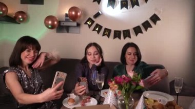 Arkadaşlar şampanya bardaklarıyla poz verip partinin şenlikli atmosferini selfie çekiyorlar. Mutlu yıllar işareti ve beyaz duvarda asılı helyum balonları