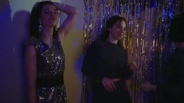 Kadınlar yüksek sesle müzik eşliğinde dans eder. Mavi ışıklar altında parlak bir arka planda doğum günü kutlaması yaparlar.
