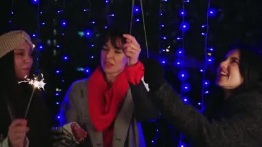 Yeni yıl arifesinde mavi ışıklara karşı maytap tutan neşeli üç kadın. Yeni yıl kutlamaları için neşeli ve parlak bir atmosfer.