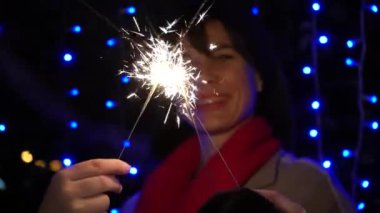 Neşeli bir gülümsemesi olan esmer bir kız önünde maytap tutuyor, video yakın çekim. Çelenklerin mavi ışıklarının arka planında neşeli bir atmosfer taşıyorum.