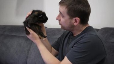 Avrupalı görünümlü bir adam tüylü hayvanını yüzüne getiriyor. Videoda bir insan ve bir kobay arasında geçen sevimli bir sahne var. Evcil hayvanların bakımı ve sevgisi için ideal.