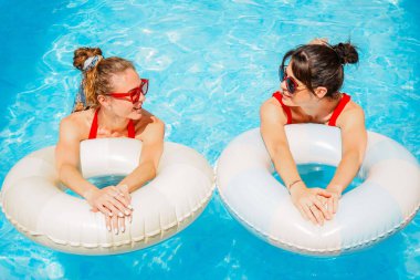 Havuz kenarında güneşli bir günün tadını çıkaran şık kırmızı mayo ve güneş gözlüklü iki kadını sersemletiyor. Şişme yüzüklü kadınlar havuzda dinleniyor.