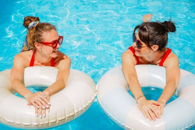 Havuz kenarında güneşli bir günün tadını çıkaran şık kırmızı mayo ve güneş gözlüklü iki kadını sersemletiyor. Şişme yüzüklü kadınlar havuzda dinleniyor.
