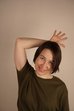 Çekici bir kadın, gri bir arka planda, bataklık tişörtü içinde, ellerini arkasına gizliyor, elini saçlarının üzerine koyuyor, aşağıya bakıyor.