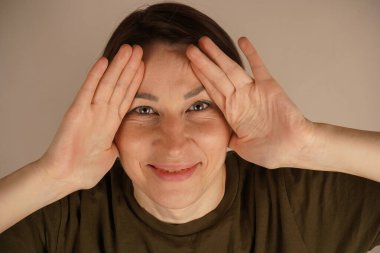 Yeşil tişörtlü, gülümseyen elleri ve yanakları izole edilmiş gri arka planda olan zeki genç bayanın fotoğrafı.