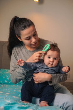 Gülümseyen genç anne kıllı fırçayla bebek saçlarını tarıyor. Yeni doğan bebeğine bakıyor. Beşik pisliği ve bebekler kepek konsepti