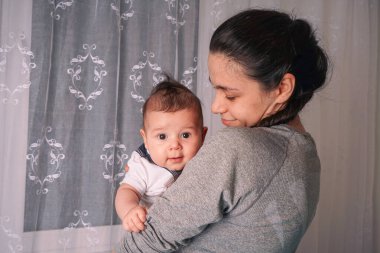 Güzel genç bir anne ayakta duruyor ve bebeğini kollarına alıyor. Evinin penceresinin önünde.