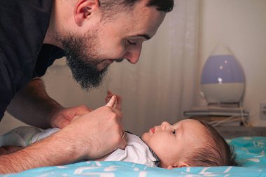 Şefkatli bir baba bebeğinin üzerine eğilir. Yumuşak mavi bir battaniyeye uzanır. Gözlerinde saf sevgi parıldar.