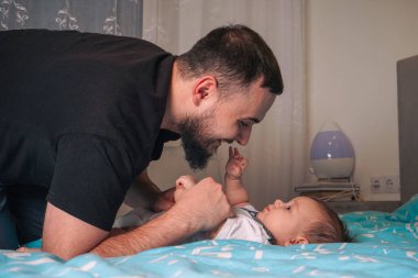 Beyaz tulum giymiş bebek, küçük kollarını babasına doğru uzatarak bir bağ kuruyor.