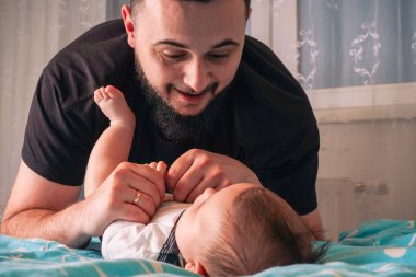 Bebek mutlu bir şekilde beyaz tulumunu tekmeliyor ve bir babanın sıcaklığına tepki veriyor.