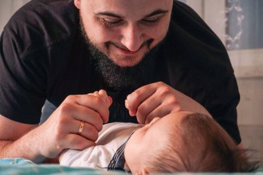 Bir baba, bebeğiyle etkileşim kurmak için eğilir. Çocuğun yumuşak beyaz tulumuyla mutlu bir şekilde öterken gülümser.