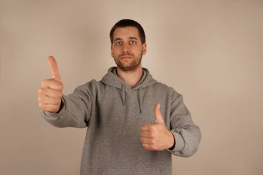 İki elini de kaldırdığında, adam mutlu bir hava yayarak baş parmağını kaldırır. Avrupalı özellikleri ve koyu renk saçları günlük gri kapüşonlusunu tamamlıyor, rahat ama şık bir görünüm sağlıyor.