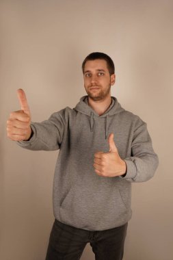 İki elini de kaldırdığında, adam mutlu bir hava yayarak baş parmağını kaldırır. Avrupalı özellikleri ve koyu renk saçları günlük gri kapüşonlusunu tamamlıyor, rahat ama şık bir görünüm sağlıyor.