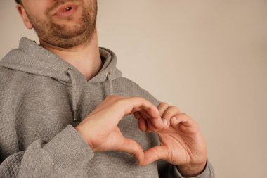 Hafif sakallı genç bir adam parmaklarıyla kalp hareketi yapar. Gri bir kapüşonlu giyiyordu, tarafsız bir arkaplanda duruyordu. Onun ifadesi nezaket ve sıcaklık yayıyor.