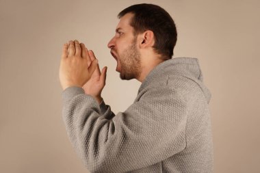 Koyu renk saçları ve canlı bir ifadeyle, adam megafonu andıran iki elini de ağzının üstüne koyuyor. Hareketleri yüksek sesle bir duyuru olduğunu gösteriyor. Görüntü heyecan ve enerji iletir.