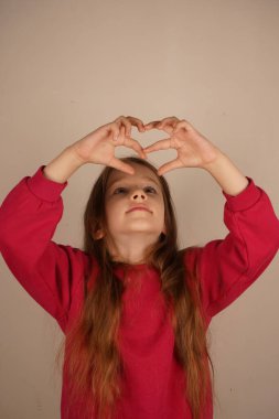 Küçük bir kız elleriyle sevgi ve şefkat gösteren bir kalp şekli oluşturur. Onun jesti sıcaklığı ve sevgiyi iletir. Nazikçe gülümser, saf duygu ve nezaket saçar.