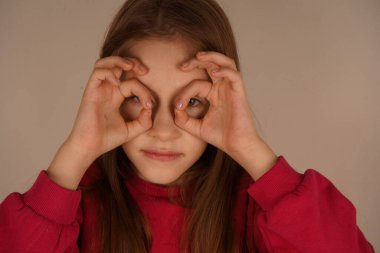 Küçük bir kız gözünün yanında gözlük taklidi yaparak parmaklarını iyi bir şekle sokuyor. Neşeli ifadesi neşe ve yaratıcılık yayıyor. Gençliğin büyüleyici bir hayal gücü.