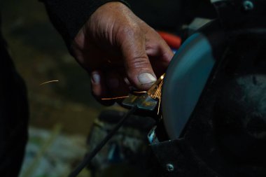 Bir zanaatkârın eli, öğütme çarkında metal bir aleti bilerken yakın plan çekimi. Sürtünme parlak kıvılcımlar yaratır. Atölyenin loş arka planı sanayi sürecini vurguluyor