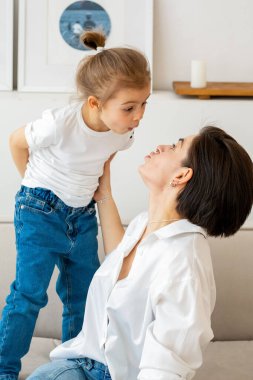 Kanepede, bir anne ve kız hassas ve neşeli bir anın tadını çıkarıyorlar. Beyaz gömlekli, inci kolyeli kadın, kızının burnuna dokunuyor, tatlı bir gülümseme uyandırıyor.