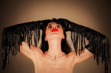 Parlak kırmızı rujlu ve püsküllü siyah deri eldivenli bir kadın portresi. Kadın siyah saçlarını tutarak iki elini de başının üstünde tutuyor.