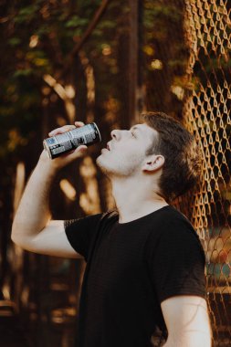Karanlık giysiler içinde, bir adam metal bir kutudan su içiyor, günlük hayatın basitliğini somutlaştırıyor. Sahne, özgünlükle dolu sıradan ve gösterişsiz bir anı yansıtıyor.