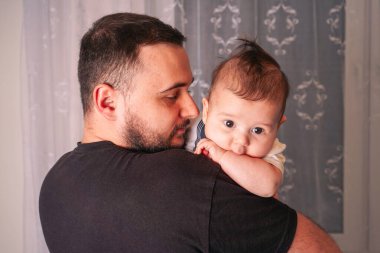Bir baba bebeğini yakın tutar, bebek merakla omzunun üzerinden bakar ve geniş gözlerle ona bakar.