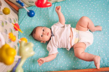 Çocuk, gözleri merakla sonuna kadar açık, mavi bir battaniyenin üzerinde yatıyor, başının üzerinde yavaşça dönen oyuncaklardan büyülenmiş.