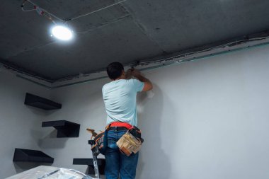 Merdivenin üzerinde duran bir adam tavanı deliyor, odaklanıyor. İş kıyafetleri içinde, yukarıdaki bölgeyi incelerken matkabı sıkıca tutuyor.