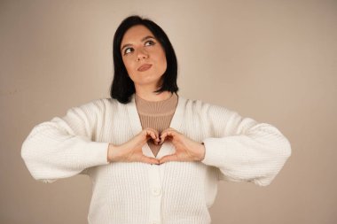 Nazikçe gülümseyen kadın elleriyle bir kalp hareketi yapar, özen ve bağlantıyı sembolize eder. Onun rahat duruşu sakinliği ve samimiyetini ifade ediyor. Bu jest gözlerinin önünde bir kalp oluşturur.