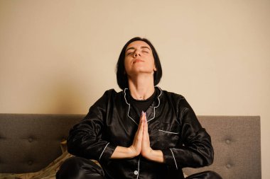 Yatakta siyah pijamalı bir kız. Lotus pozisyonunda bir kadın sabah meditasyon yapıyor. Yatakta ayin yapan bir kadın. Elleri avuç içlerine katlanmış. Şafağın loş ışığı