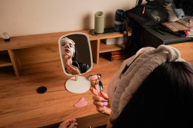 Saç bandı ve siyah ipek pijama giyen bir kadın nemlendirici bir maskeyle cildine bakar. Masadaki küçük bir ayna onun parlak cildini yansıtıyor. Masa aynasında yansıyan bir kız.