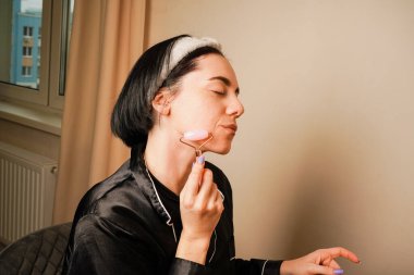 Yüz silindiri kullanan bir kadın çene hattına masaj yapar. Rahatlatıcı bir cilt bakım rutini. Doğal güzelliği ve zarif küpeleri sofistike sahneyi tamamlıyor. Işıklar huzurlu bir ortam yaratıyor.