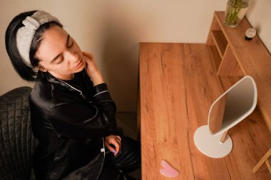 Bir kadın, çene hattına masaj yapmak için narin bir metal silindir kullanır. Siyah pijamalı bir kız aynanın önündeki masada oturuyor. Yüzündeki ifade kendini önemsemeyi yansıtıyor.