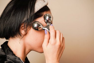 Bir kadın evinde dinleniyor, bir silindirle kendine yüz masajı yapıyor. Siyah saçları ve siyah pijamaları sahneyi vurguluyor. Odak noktası onun parlak, sağlıklı teni.