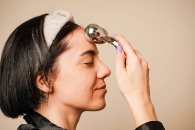 Bir kadın evinde dinleniyor, bir silindirle kendine yüz masajı yapıyor. Siyah saçları ve siyah pijamaları sahneyi vurguluyor. Odak noktası onun parlak, sağlıklı teni.