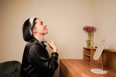 Bir kadın, çene hattına masaj yapmak için narin bir metal silindir kullanır. Siyah pijamalı bir kız aynanın önündeki masada oturuyor. Yüzündeki ifade kendini önemsemeyi yansıtıyor.