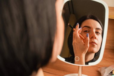 Alnına rimel süren bir kadın, aynadaki yansımasına hayran kalır. Onun kısa siyah saçı görünüşüne zarafet katıyor. Oda sıcak ahşap tonlarla vurgulanıyor.