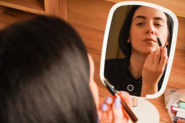 Alnına rimel süren bir kadın, aynadaki yansımasına hayran kalır. Onun kısa siyah saçı görünüşüne zarafet katıyor. Oda sıcak ahşap tonlarla vurgulanıyor.