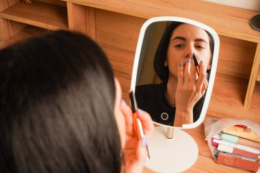 Makyaj yapan bir kadının yakın çekimi dudaklarını kapladığını gösteriyor. Onun gevşemiş ifadesi kendini korumayı yansıtıyor. Arkasında, ahşap bir raf ve küçük dekoratif eşyalar rahat bir atmosfer yaratıyor.