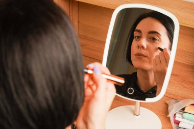 Makyaj yapan bir kadının yakın çekimi dudaklarını kapladığını gösteriyor. Onun gevşemiş ifadesi kendini korumayı yansıtıyor. Arkasında, ahşap bir raf ve küçük dekoratif eşyalar rahat bir atmosfer yaratıyor.