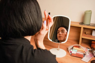 Genç bir kız küçük bir aynanın yanında oturuyor. Kızın yanına makyaj malzemesi sürme özelliği. Aynada, kısa siyah saçlı bir kızın yansıması.