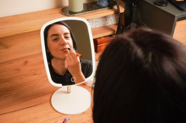 Makyaj yapan bir kadının yakın çekimi dudaklarını kapladığını gösteriyor. Onun gevşemiş ifadesi kendini korumayı yansıtıyor. Arkasında, ahşap bir raf ve küçük dekoratif eşyalar rahat bir atmosfer yaratıyor.