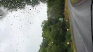 Arabanın camında yağmur damlaları dikiz aynasına odaklanmış, dışarıda fırtınalı bir atmosfer, yağmurlu yolculuk konsepti, kötü hava koşullarında araç güvenliği. Yan ayna yağmurlu araba penceresinden geçen yolu yansıtıyor, bulutlu gökyüzünü.