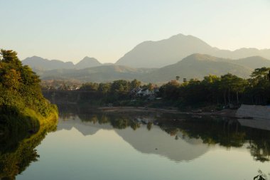 Gün batımında dağların, ormanların ve siluetlerin manzarası, Luang Prabang, Laos 'tan geçerken Nam Khan Nehri' nin sularını yansıtıyor.