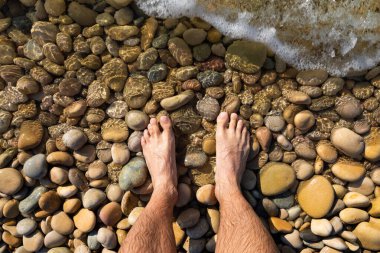 Feet of a man, from a subjective view, in the water at the seashore, on a pebble beach, washed by the waves of the Mediterranean Sea