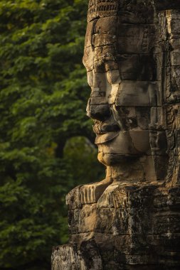 Taşa oyulmuş kocaman bir yüz, Bayon, Angkor, Kamboçya tapınağına gelen ziyaretçileri izliyor, arka planda yeşil bir orman duvarı var.