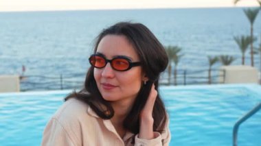 Genç ve çekici bir kızın yakın plan portresi yüzme havuzu ve okyanusun arka planına karşı. Çekici milenyum kızı pembe güneş gözlüğü takıyor kameraya bakıyor ve gülümsüyor.. 