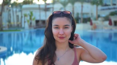  Genç ve çekici bir kızın yakın plan portresi yüzme havuzunun arka planına karşı. Çekici milenyum kızı pembe güneş gözlüğü takmış kameraya bakıyor ve gülümsüyor.