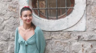 Yeşil elbiseli ve kırmızı bandanalı bir kadın pencerenin önünde duruyor. Gülümsüyor ve mutlu.